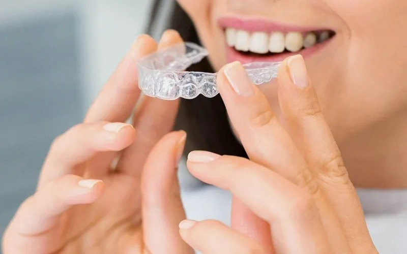 A woman smiling while holding a clear Invisalign aligner